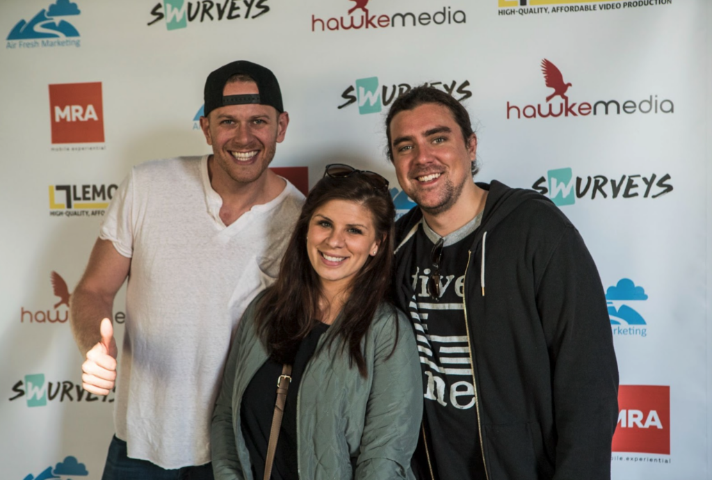 Three individuals posing together with smiles at an event with sponsor banners in the background.