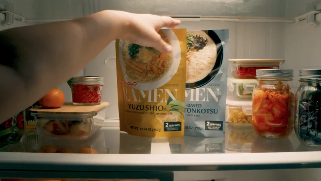 A hand reaches for a packet of Yuzu Shio ramen inside a refrigerator, next to a Tonkotsu ramen packet and jars of various pickled vegetables.