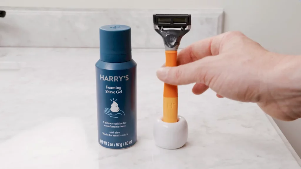 A hand holding an orange and black razor next to a blue can of Harry's Foaming Shave Gel on a white bathroom countertop.
