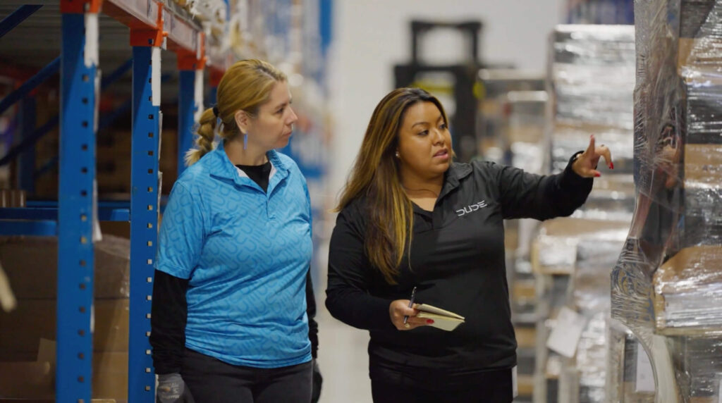 Two women stand in a warehouse aisle; one points toward shelves while the other looks on attentively, holding a notepad.