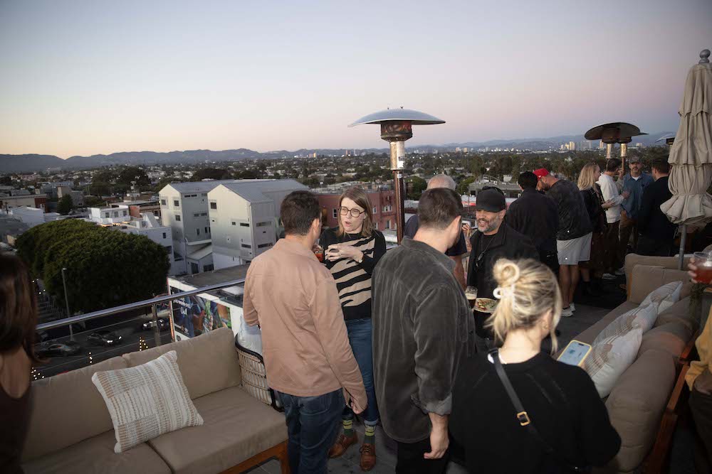 A group of people socialize on a rooftop patio at sunset during LA Tech Week, with city buildings and mountains visible in the background.