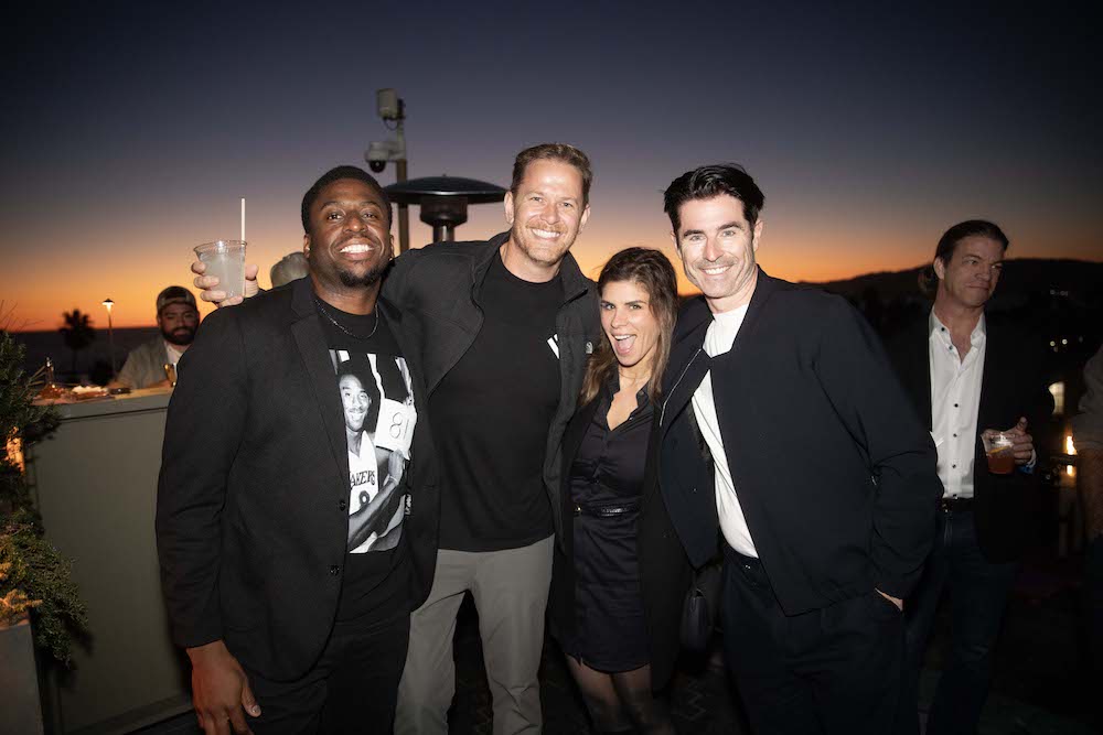 Four people pose together and smile at an outdoor LA Tech Week event during sunset, with other guests and lights visible in the background.