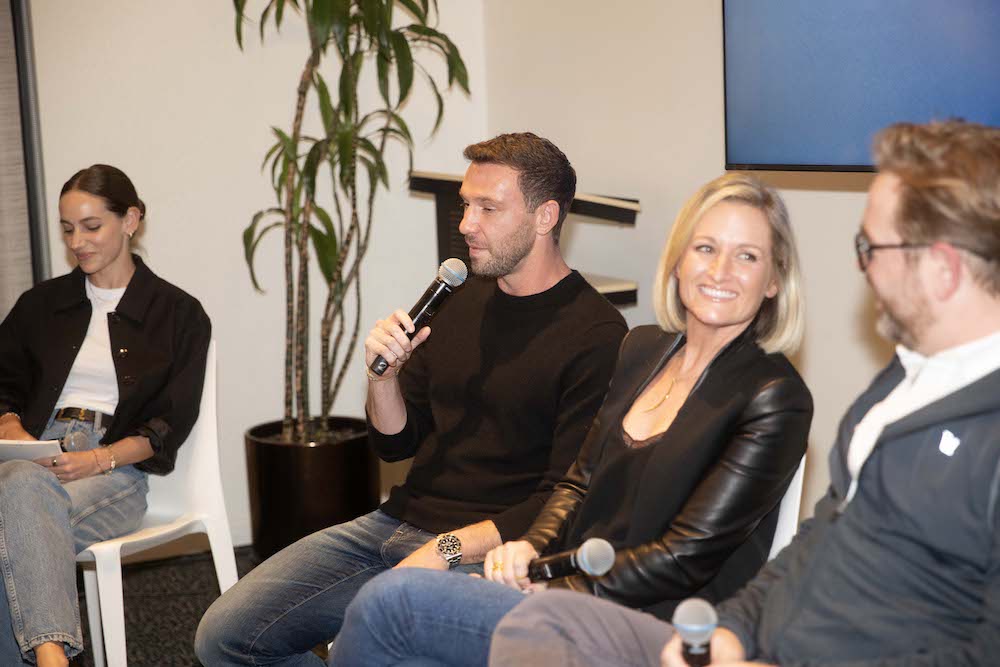 Four people sit indoors during LA Tech Week; one man speaks into a microphone while the others listen and smile. A potted plant and a TV screen are visible in the background.