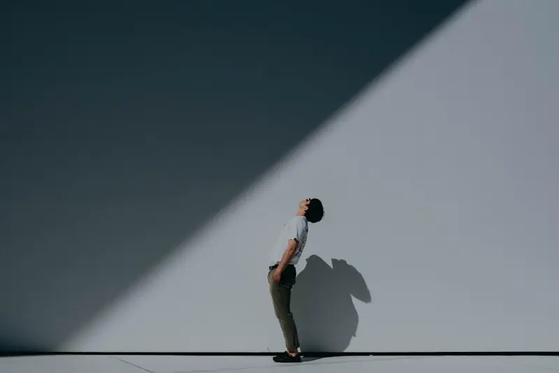 A person stands against a white wall, leaning back and looking up, with a sharp diagonal shadow cast across the wall and ground.