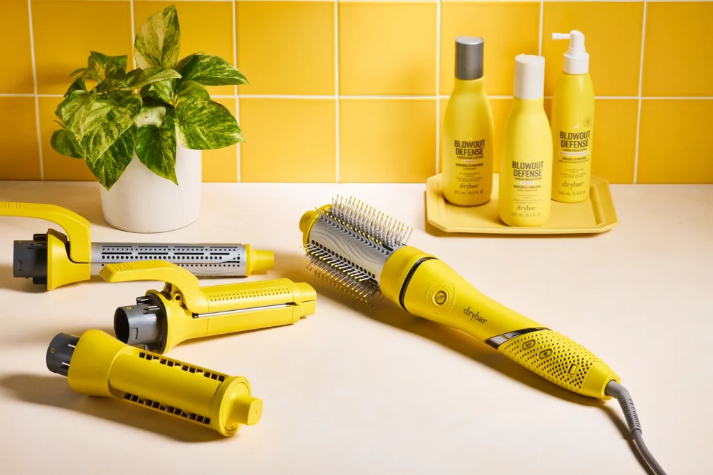 A yellow hot air brush with detachable barrels is on a counter beside plant, hair care bottles, and a yellow tiled wall.
