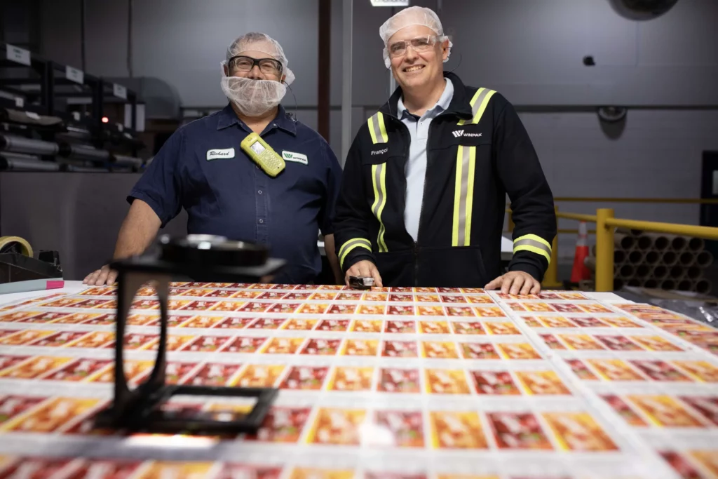 Two men in hairnets and safety glasses stand behind a table covered with printed sheets in a factory setting.