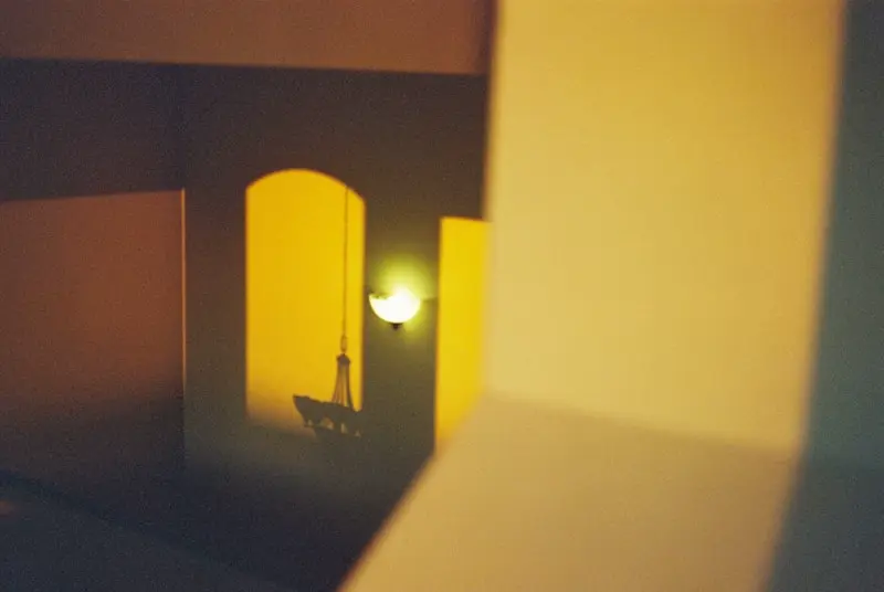 Blurred view of a warmly lit interior showing a wall sconce and chandelier through an archway, with out-of-focus architectural elements in the foreground.