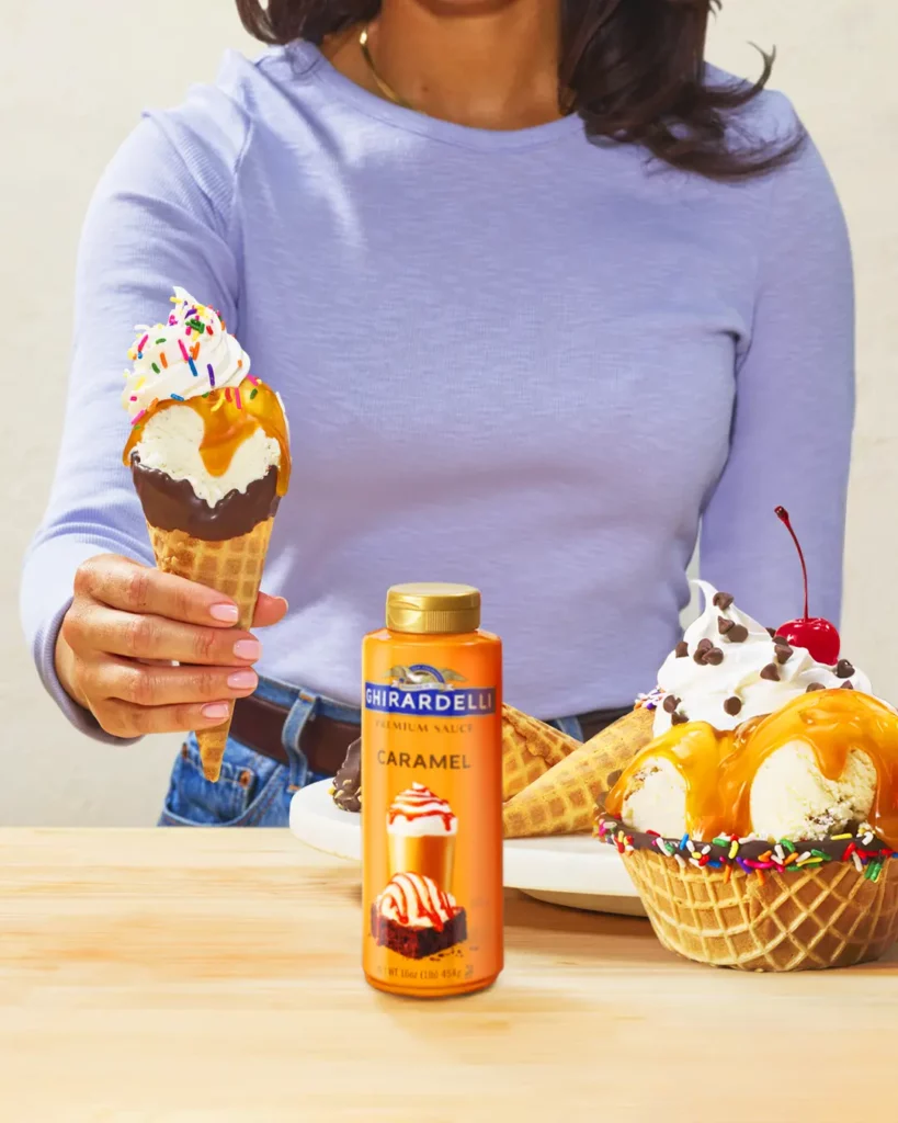 A person holding an ice cream cone with caramel sauce and sprinkles, with a bottle of Ghirardelli caramel sauce and a plate of ice cream sundaes on the table.