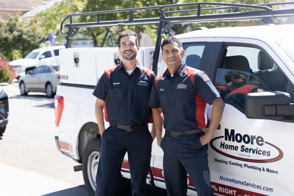 Two Moore Home Services technicians stand in front of a company truck, wearing uniforms and smiling at the camera on a sunny day.