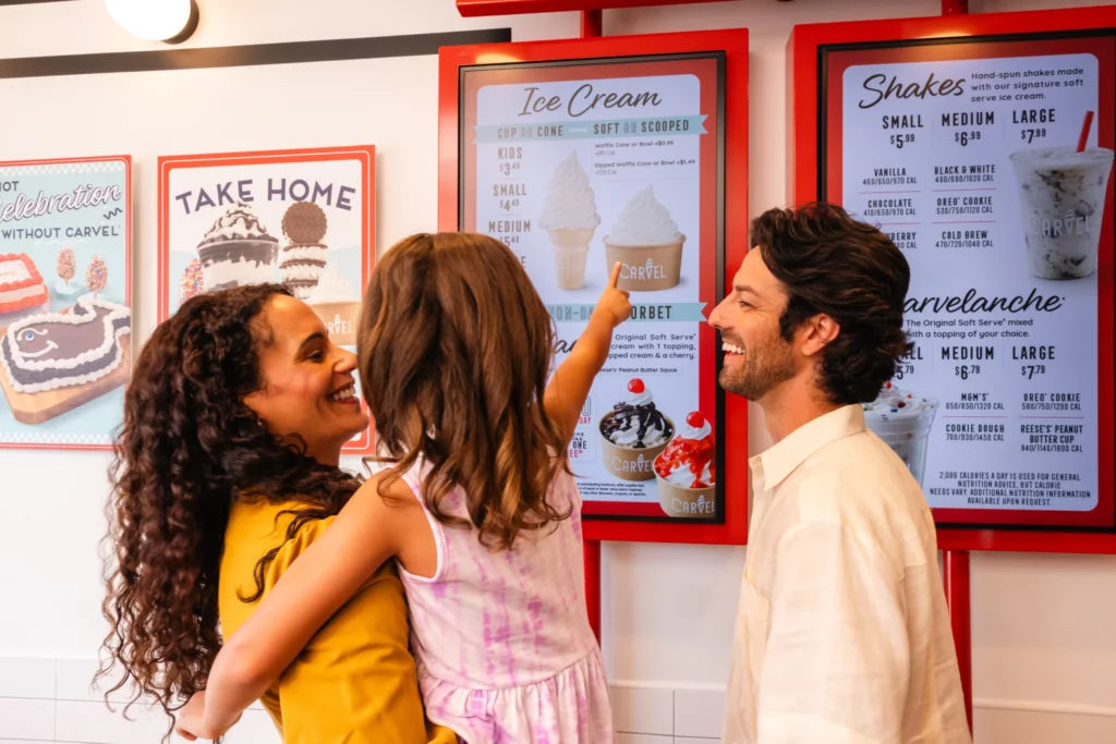 A woman holds a young girl who points at an ice cream menu on the wall while a man stands beside them, all smiling inside an ice cream shop.