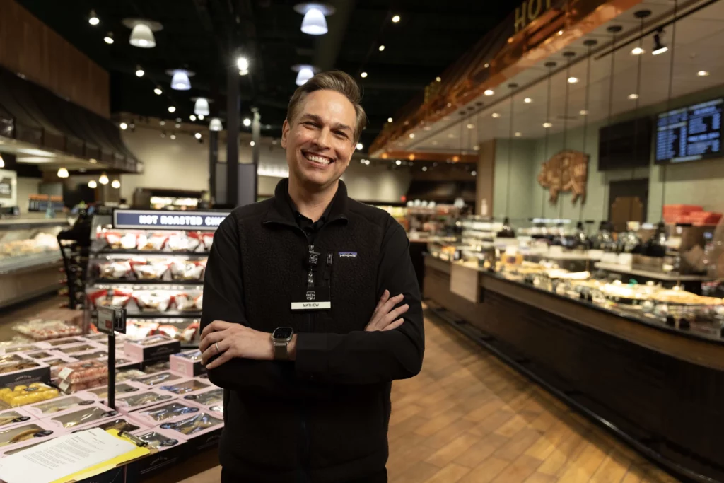 A smiling man with folded arms stands in the prepared foods section of a grocery store. Display cases with food and bright overhead lights are visible.