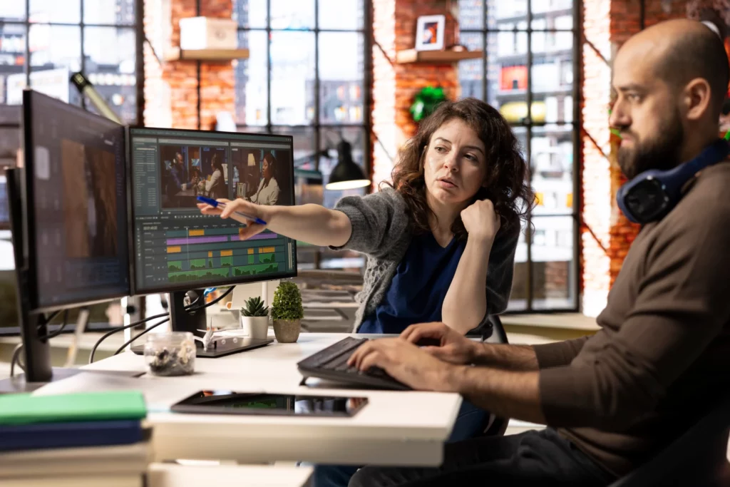 Two people work together at a desk with dual monitors displaying video editing software in a modern office with large windows.