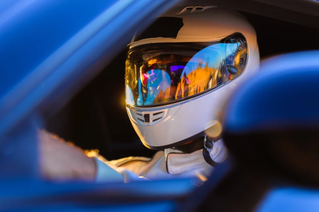 A person in a white racing helmet sits in the driver's seat of a blue car, seen through the open window, with reflections visible on the helmet's visor.