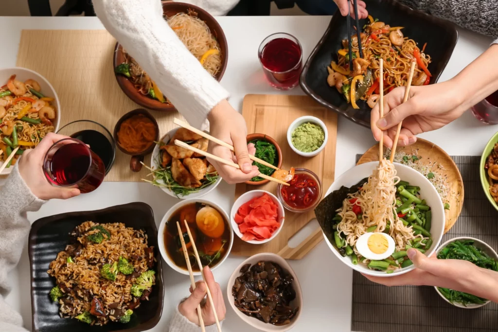 People use chopsticks to share various Asian dishes, including noodles, rice, vegetables, and sauces, arranged on a table with drinks.