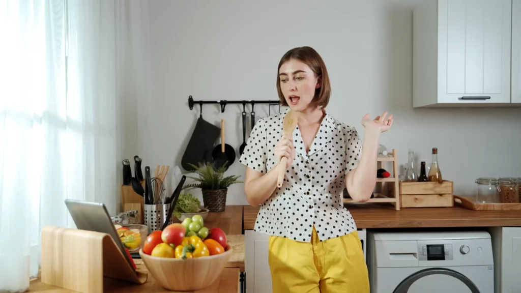 A woman in a polka dot shirt and yellow pants sings into a wooden spoon in a kitchen, with vegetables and a tablet on the counter.