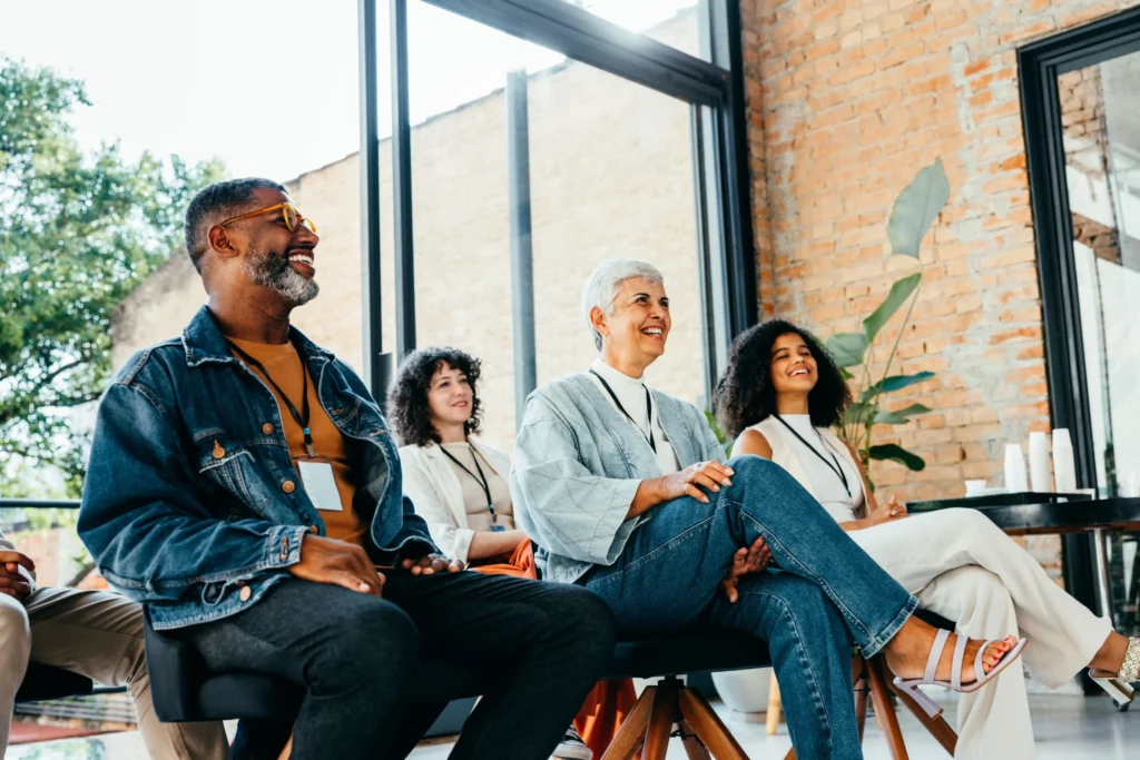 Four adults sit in chairs, smiling and attentively listening during what appears to be a group meeting or seminar in a bright, modern room with brick walls and large windows.