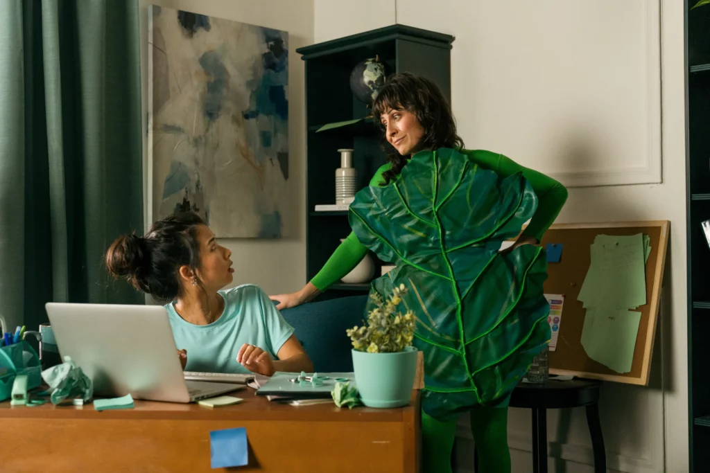 Two women in an office; one sits at a desk with a laptop, while the other stands beside her wearing a large green leaf costume.