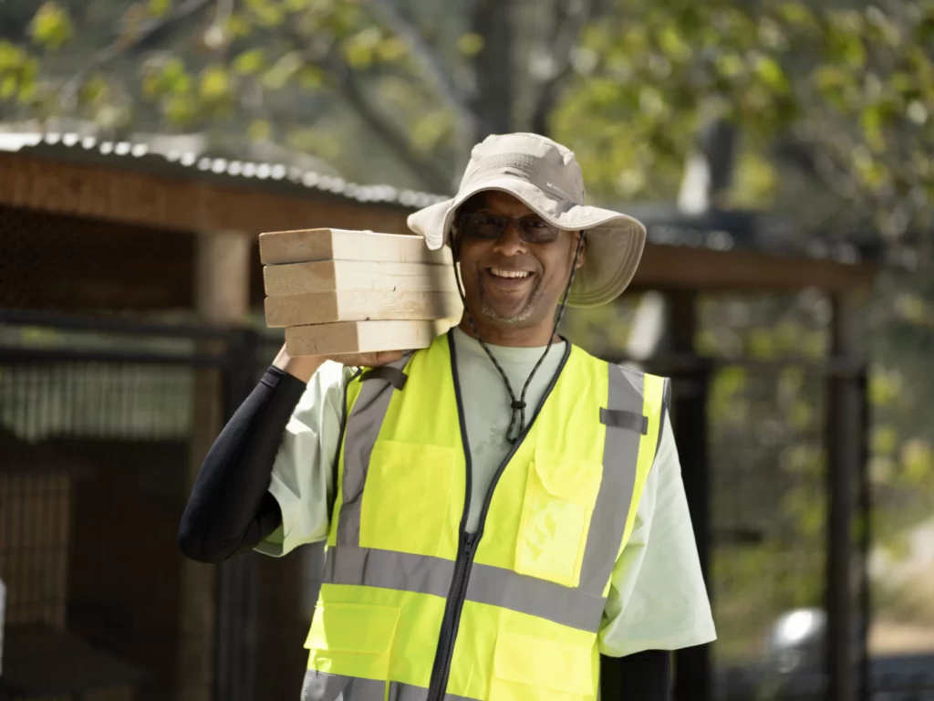 A person wearing a high-visibility vest and sun hat smiles while carrying several wooden planks on their shoulder outdoors.