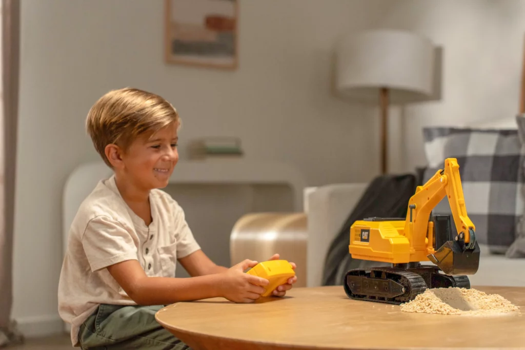 A young boy sits at a table indoors, using a remote control to operate a yellow toy excavator with a small pile of sand in front of it.