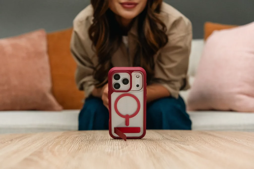 A person sits on a couch with their face out of focus, while a smartphone in a red and white MagSafe case is positioned upright on a wooden table in the foreground.