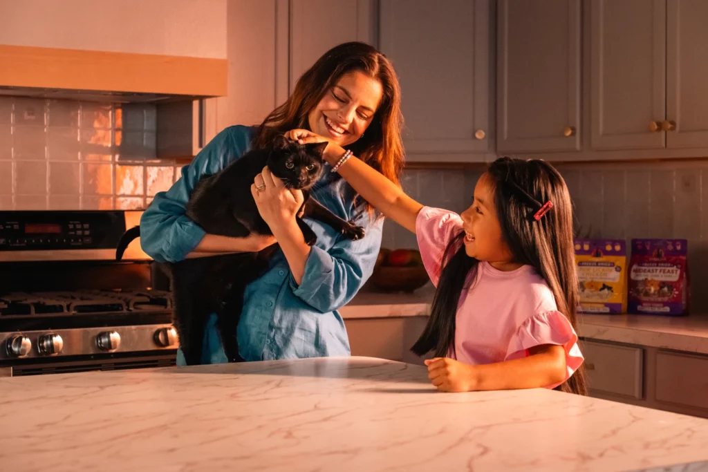 An adult holds a black cat while a child pets it at a kitchen counter; pet treat bags are visible in the background.