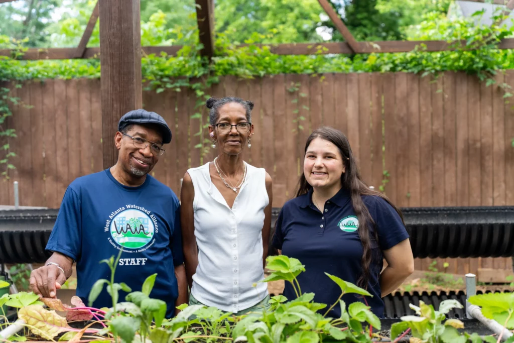 Three adults stand together in a garden, posing for a photo. Two wear shirts with a green and blue logo and the word "STAFF." Green plants and a wooden fence are visible in the background.