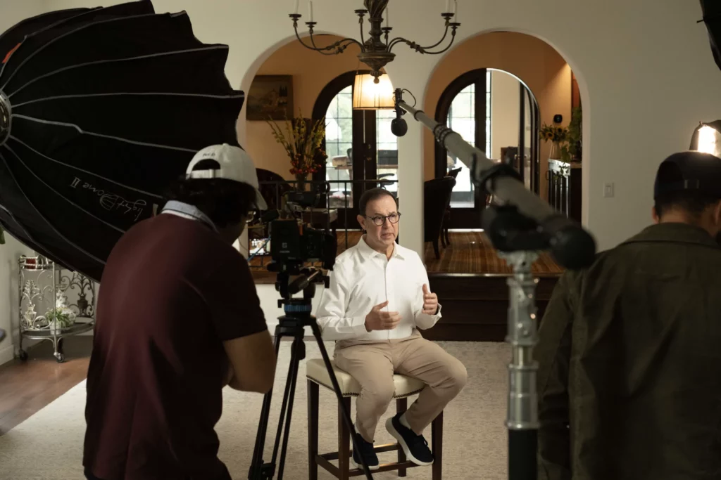 A man in a white shirt sits on a stool being filmed in a well-lit room, with a camera crew, lights, and audio equipment visible.