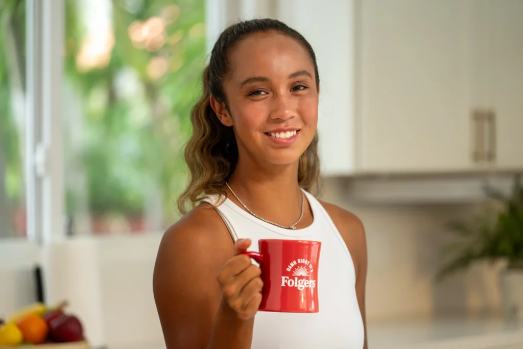 A woman in a white sleeveless top holds a red Folgers coffee mug and smiles in a bright kitchen with fruit visible in the background.