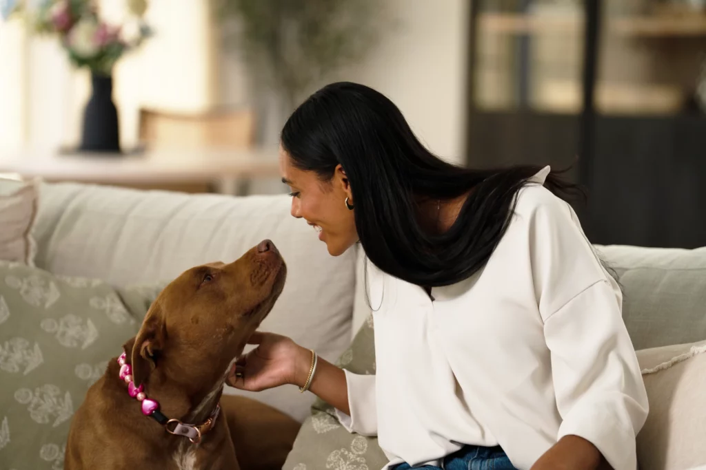 A woman sits on a couch, smiling and petting a brown dog wearing a pink collar with a heart-shaped tag.