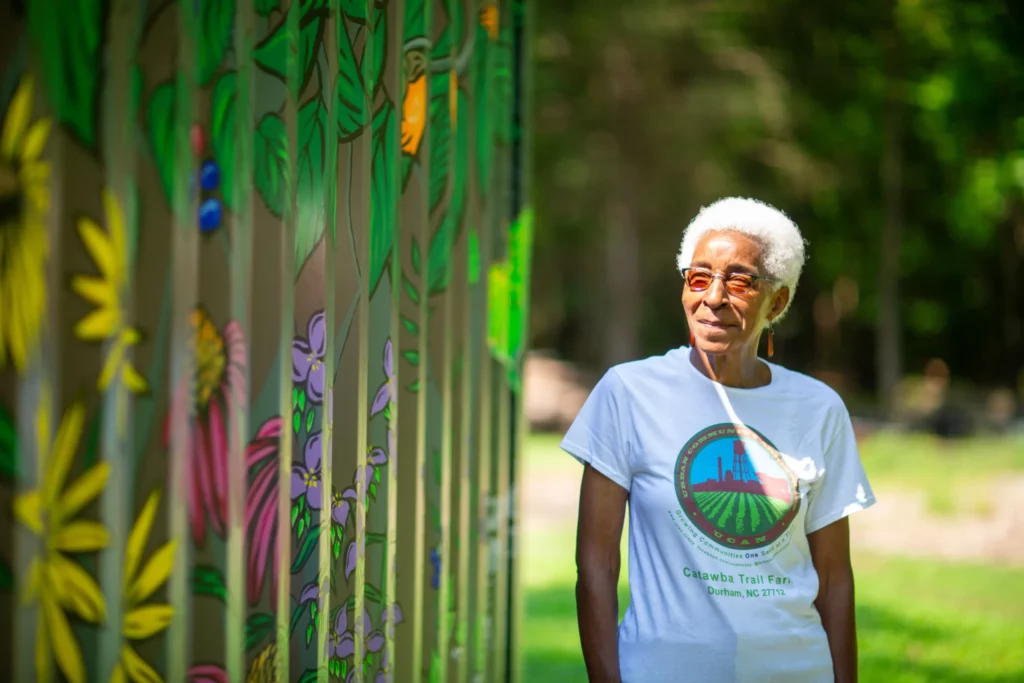 An older woman with short white hair and glasses stands outdoors near a painted wall with colorful flowers and plants, wearing a white t-shirt with a farm logo.