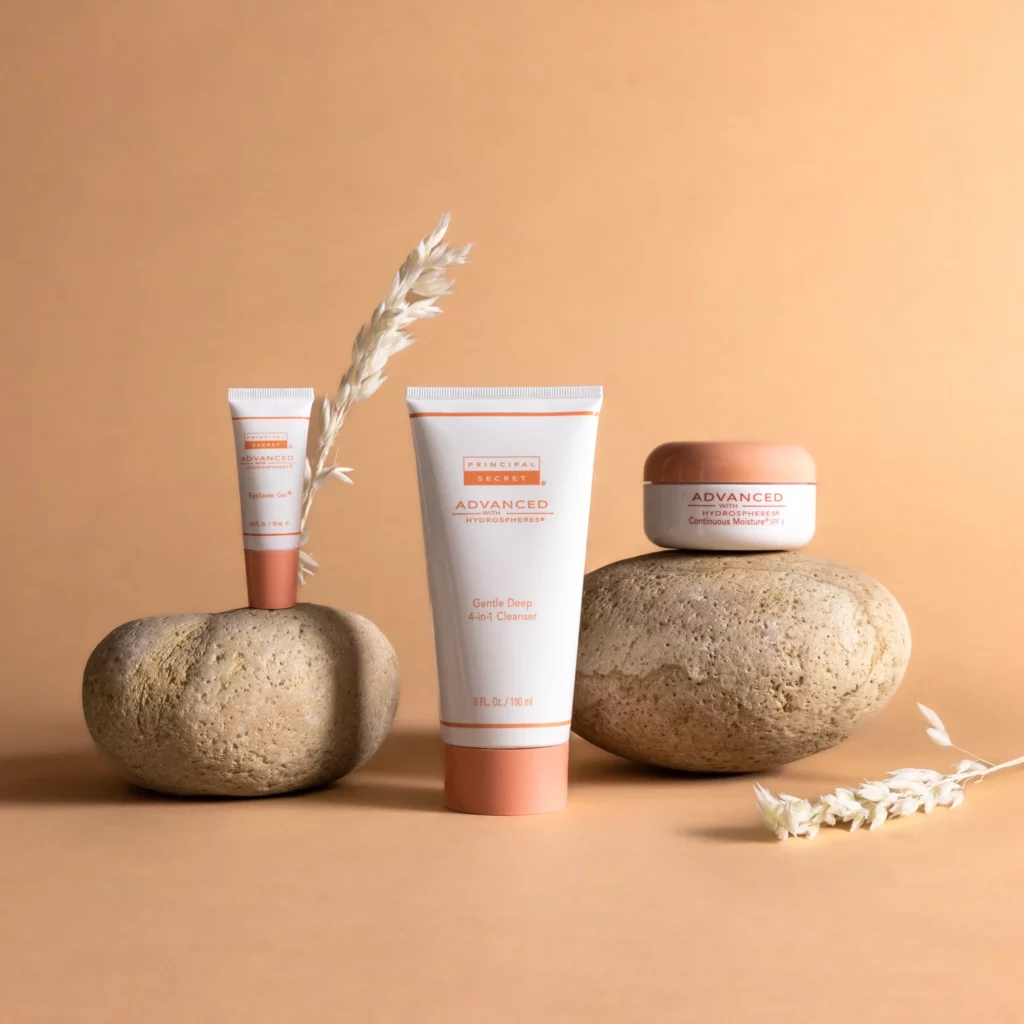 Three skincare products and some dried plants are arranged on smooth stones against a beige background.