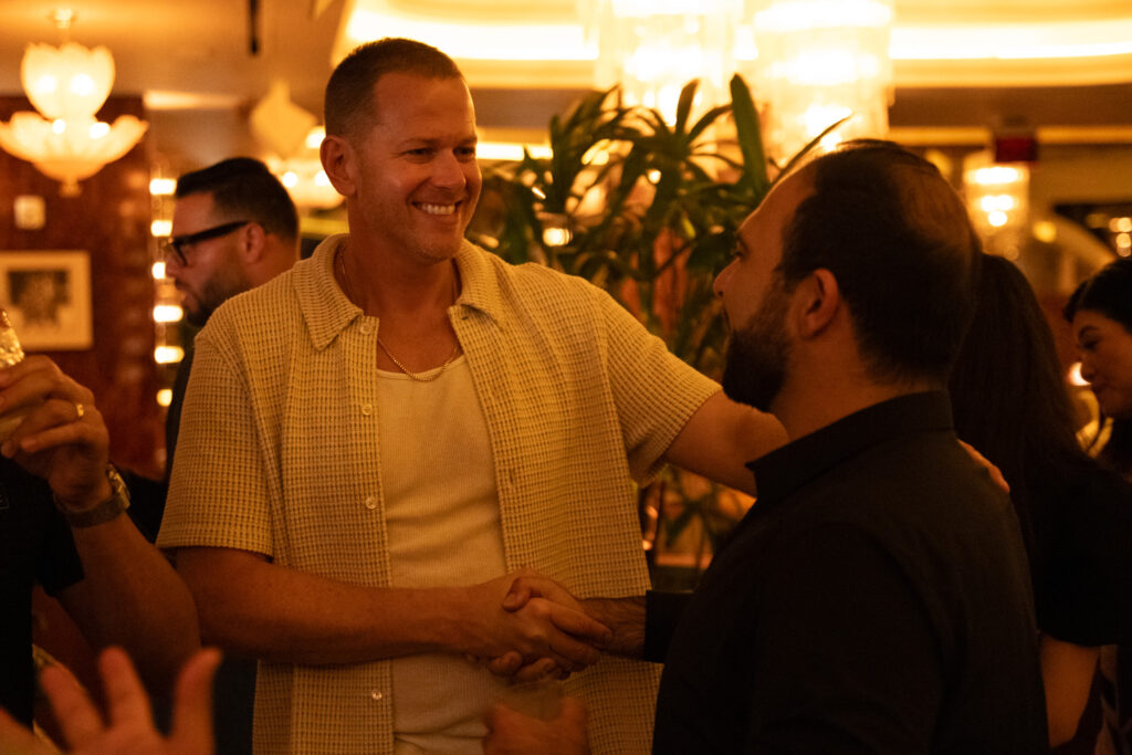 Two men shake hands and smile in a warmly lit indoor setting with other people and plants in the background.