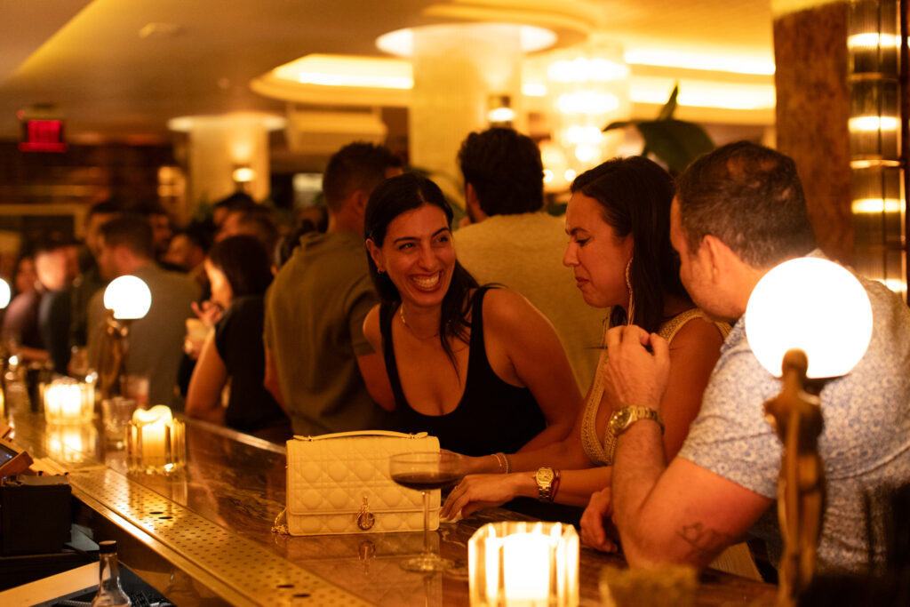 A crowded bar with people socializing; two women and a man are in focus at the counter, with warm lighting and candles creating a cozy atmosphere.