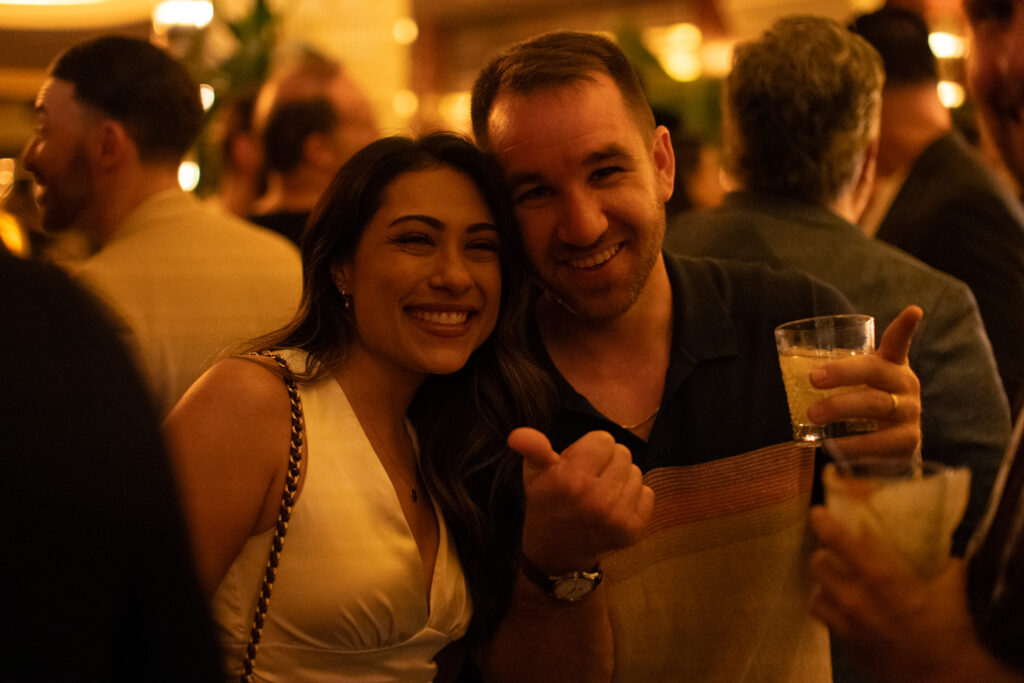 A woman in a white dress and a man holding a drink smile and pose for a photo in a dimly lit, crowded indoor setting.