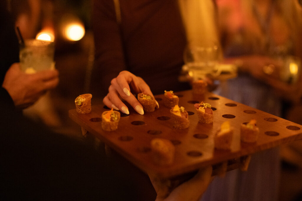 A person selects a small appetizer from a wooden serving tray with circular holes at a dimly lit gathering. Other guests and drinks are visible in the background.