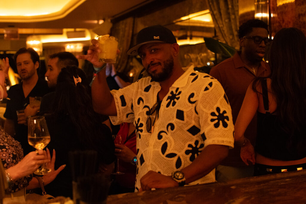 A man in a patterned shirt holds up a drink at a crowded bar, surrounded by people socializing under warm, dim lighting.