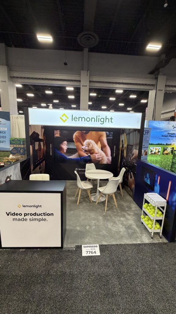Lemonlight trade show booth with a table, chairs, and video on display; a sign reads “Video production made simple,” and tennis balls are visible on the right.