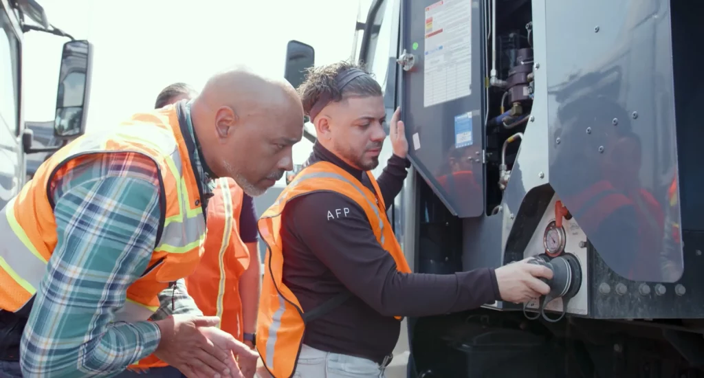 Two workers in safety vests examine the controls of a machine. One man is holding open a panel door.